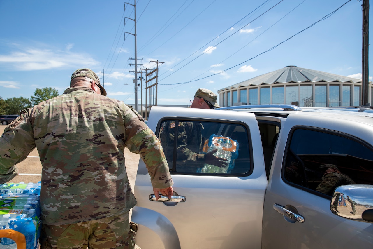 A soldier holds the door of a car open while another places a case of bottled water on the back seat.