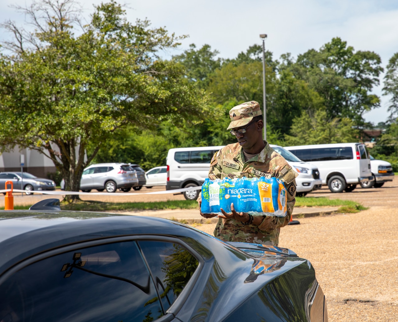 A soldier carries a case of water toward the back of a car.
