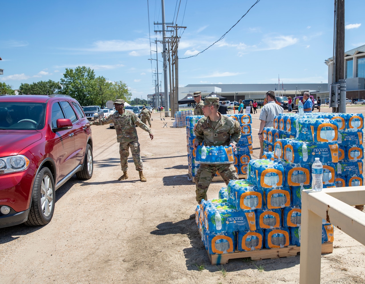 A soldier holds the door of a car while another soldier grabs a case of bottled water.