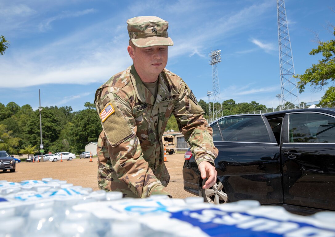 A soldier reaches for a case of water from a pallet with a car in the background.