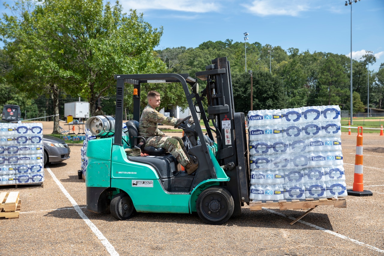 A soldier operates a small vehicle while moving pallets of water in a parking lot.