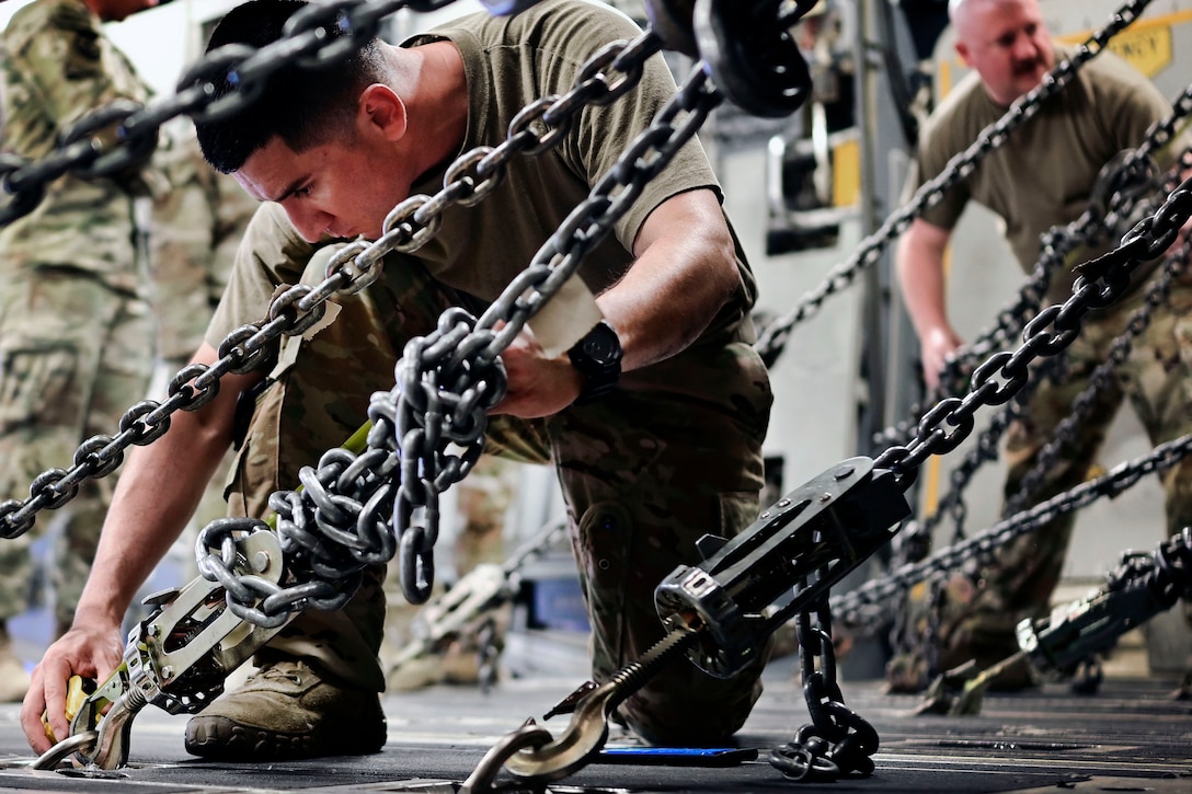 A soldier kneels while measuring a chain that is bolted to the floor.
