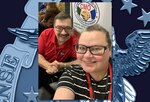 A smiling man and woman take a selfie at a conference on the expo floor. She wears a striped shirt and has her hair in a slicked back ponytail while he has on a red shirt and has a mustache. Both wear glasses.