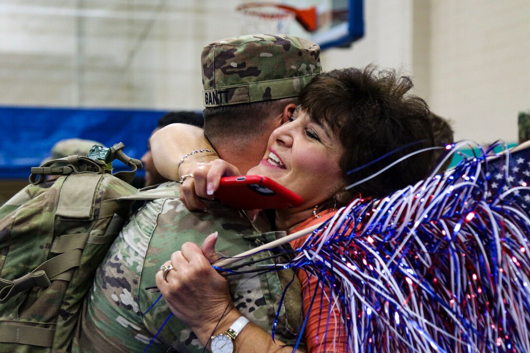 A civilian holding a flag hugs a service member in a gym.