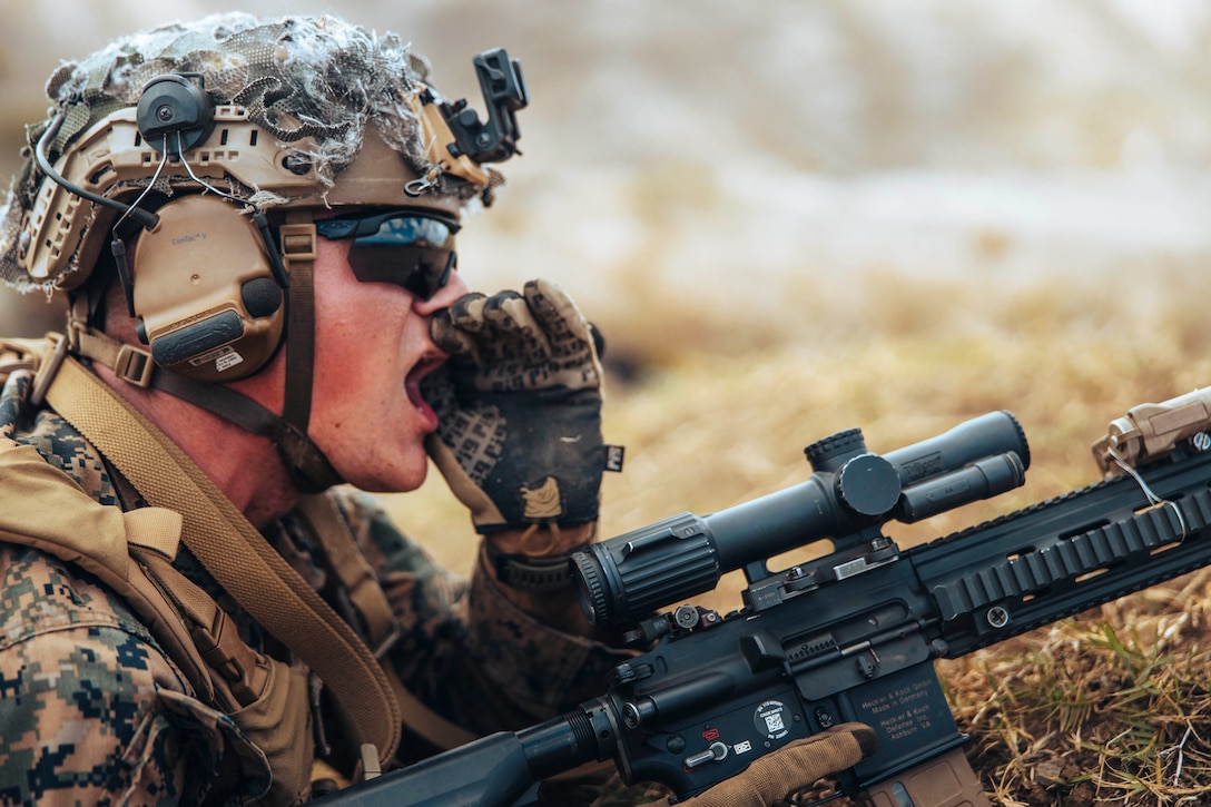 A Marine holding a weapon yells while kneeling in a field.