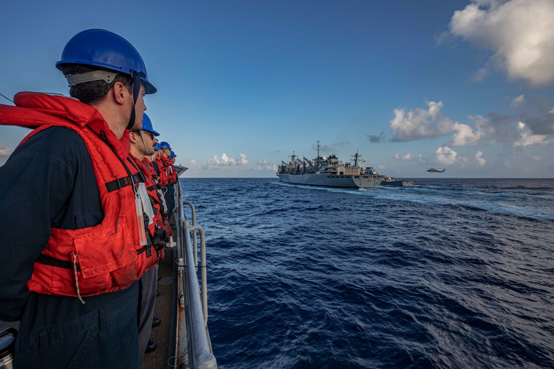 Sailors stand at the rails of a ship and look at two ships sailing side-by-side with a helicopter hovering above.