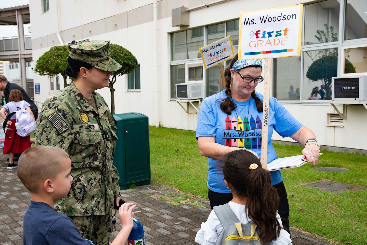 A service member and civilian stand outside a school with kids.