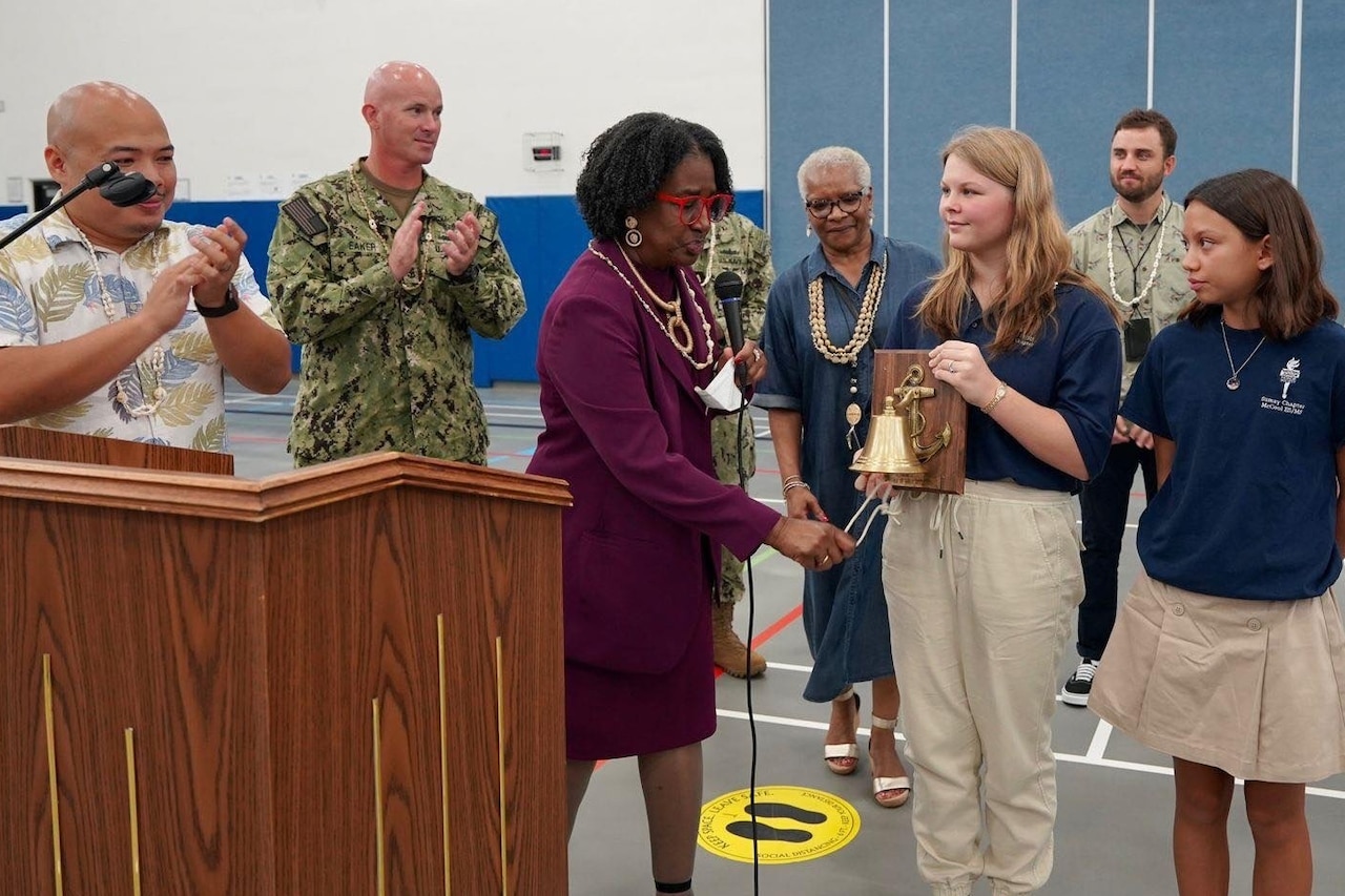 A student holds up a bell as military and civilian adults applaud in a gym-type room.