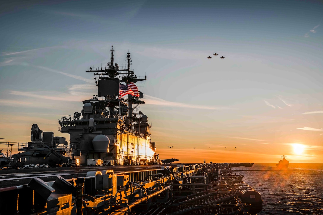 Aircraft fly above two ships traveling through waters; the sun can be seen in the background.