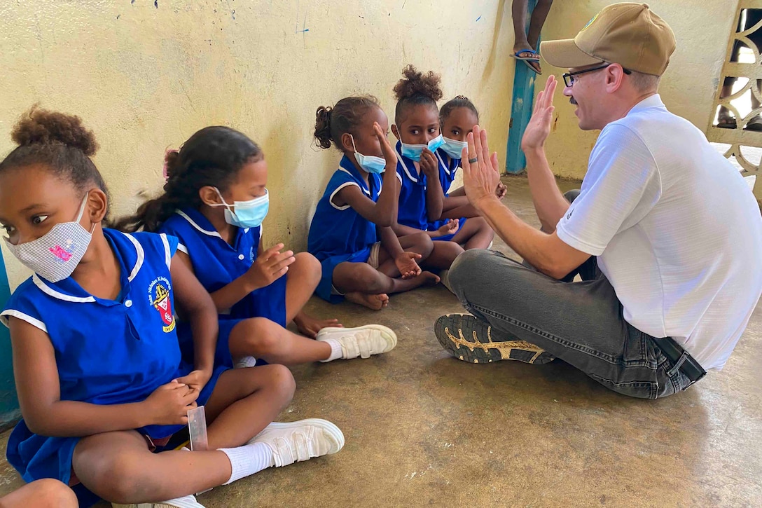 A sailor sits on the floor clapping hands with children.