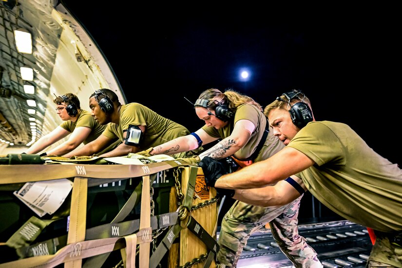 Four airmen wearing headphones push pallets onto an aircraft.