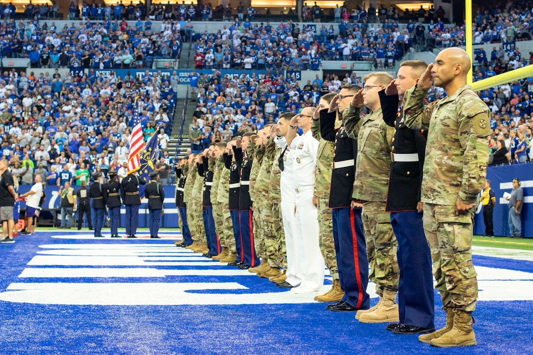 Service members lining the end zone of a football field salute an American flag with hundreds of fans watching.