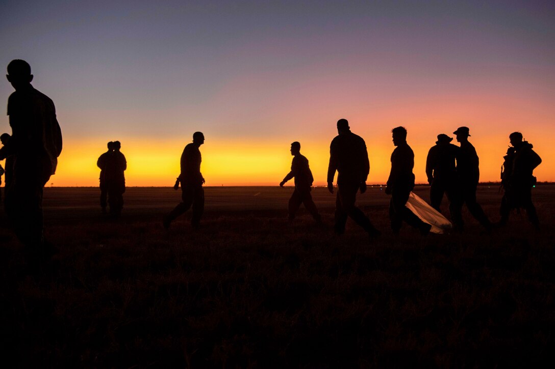 Marines walk on a flightline under a sunlit sky as seen in silhouette.