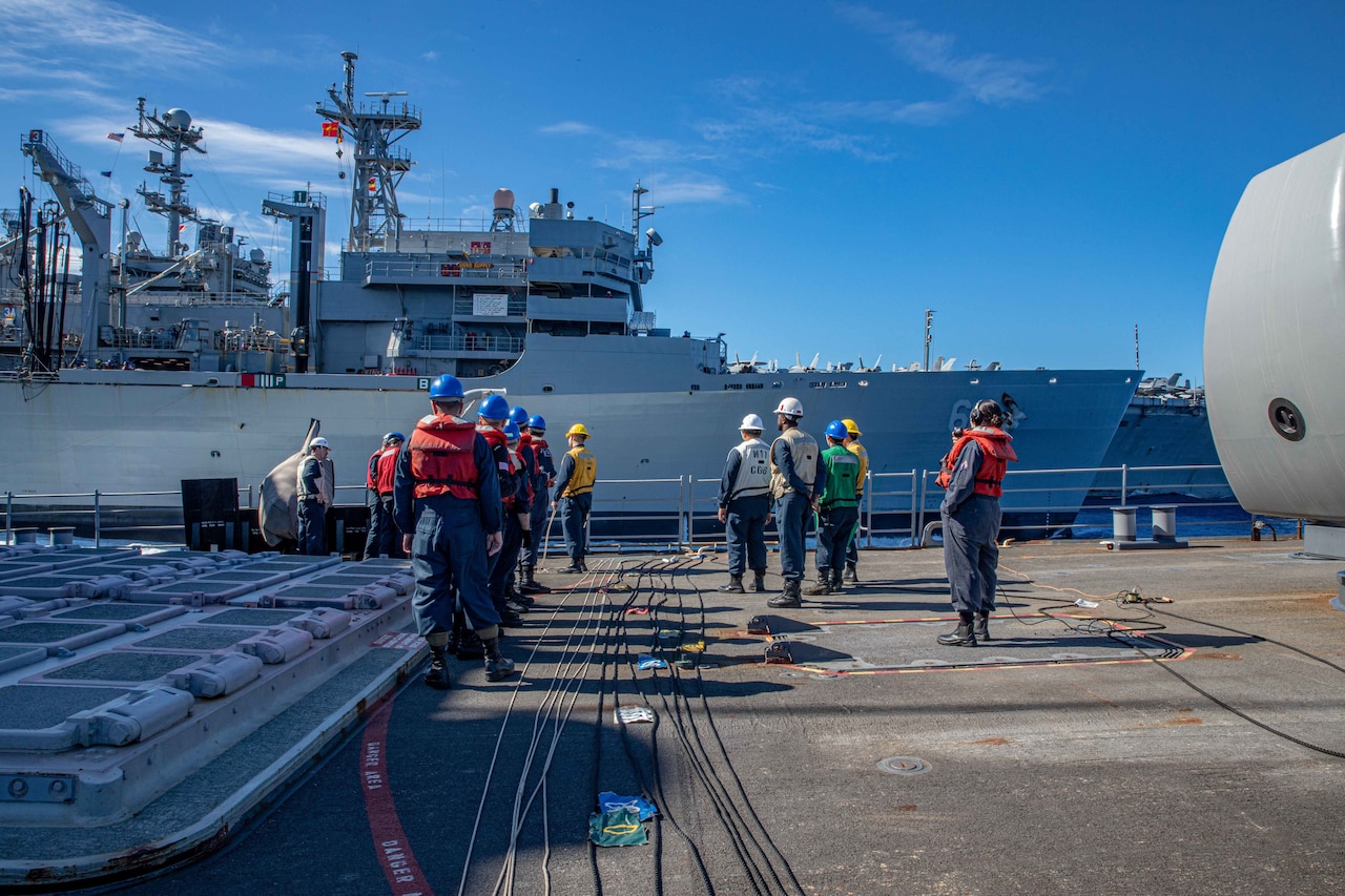 Personnel stand near line laying on the deck of a ship and look at another ship sailing at their side.