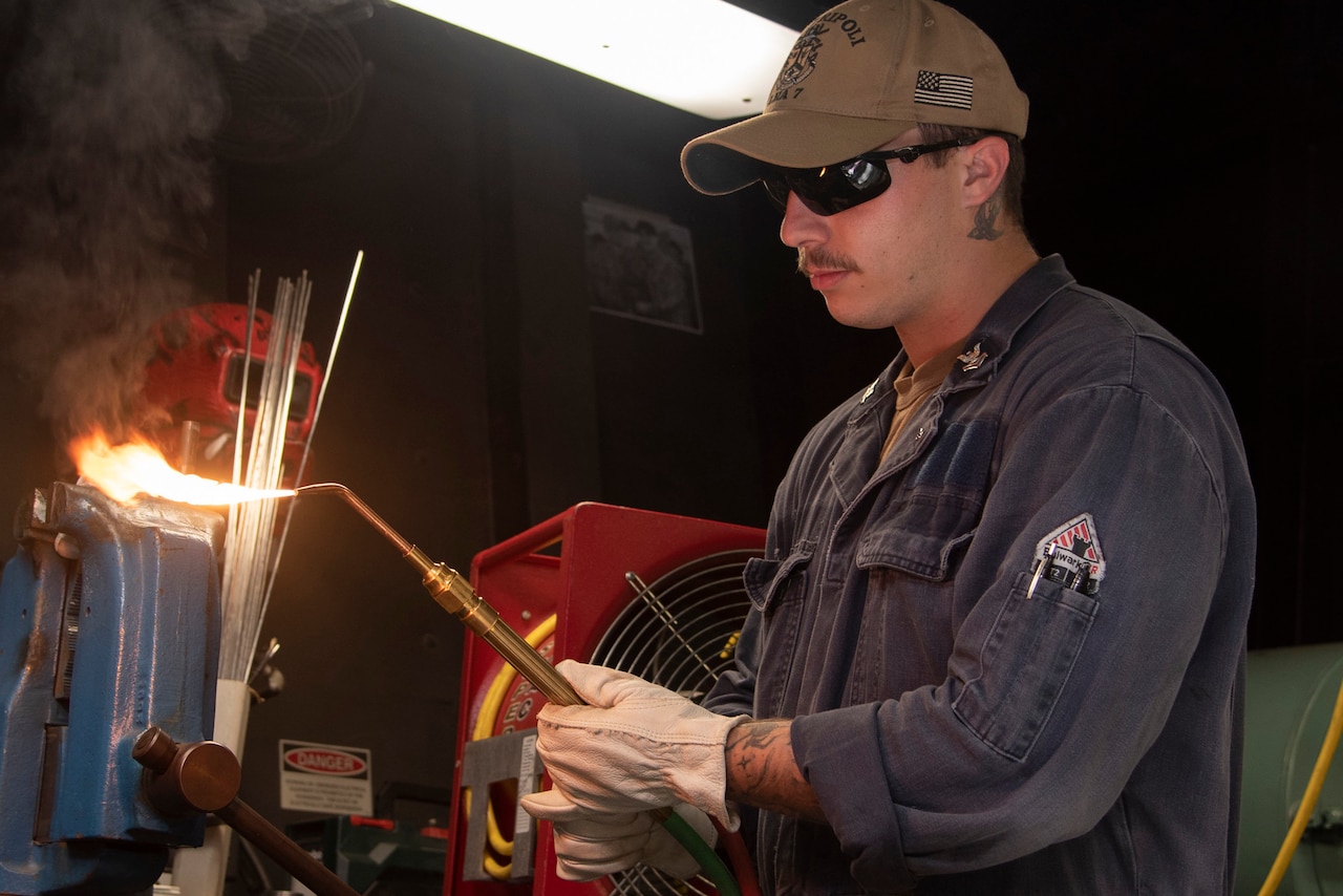 A sailor wearing gloves holds a lit brazing torch.