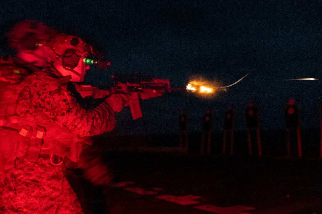 A Marine in red light fires a  weapon creating sparks at night from the deck of a ship.