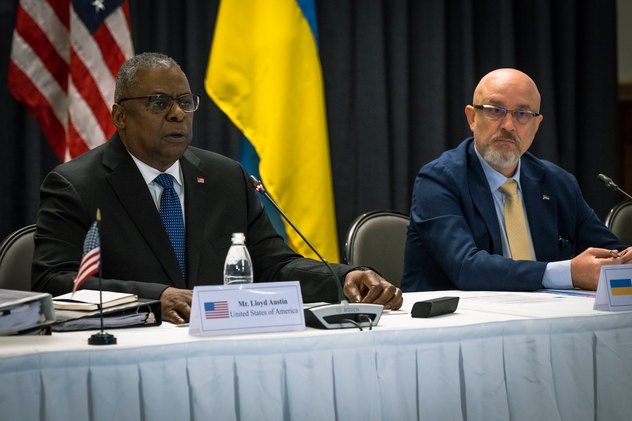 Two men sit behind a long table as one makes remarks.