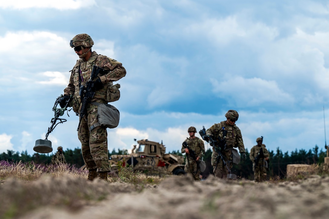 Soldiers walk through a grass field while using handheld mine detectors.