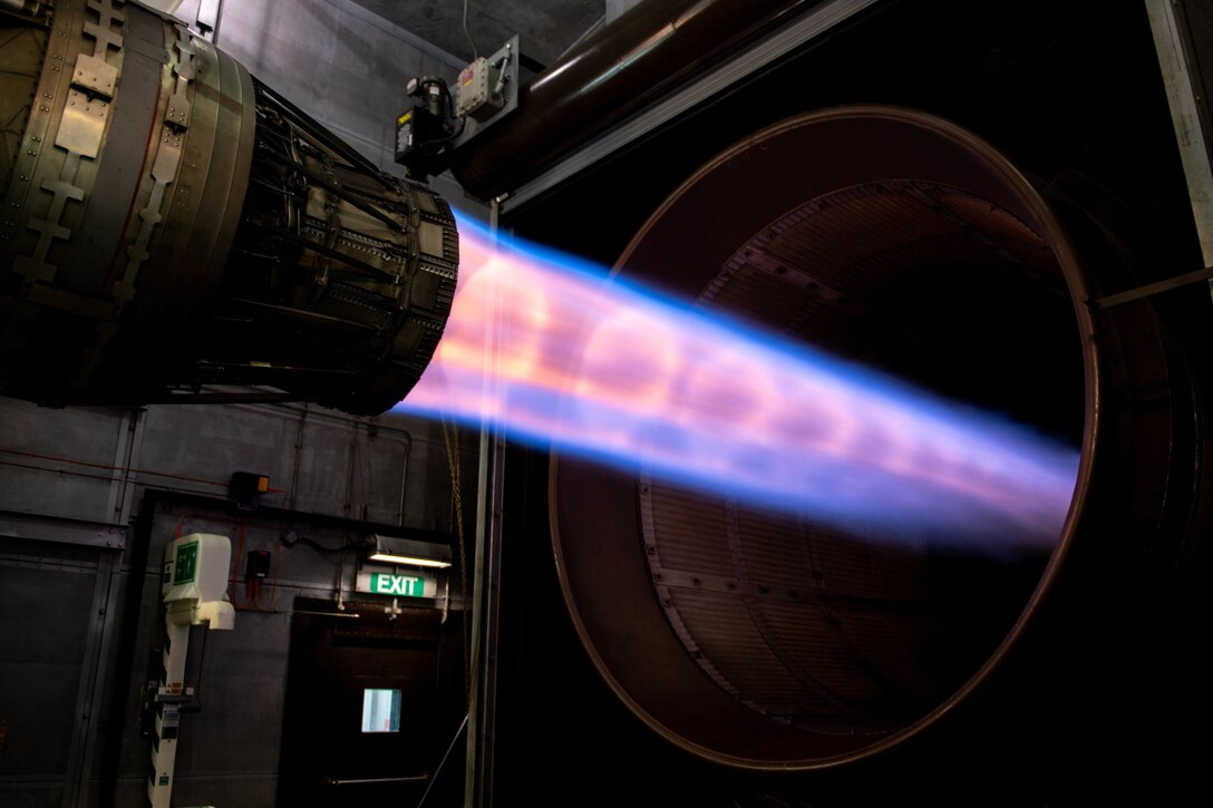 A ray of light-colored gas shoots from a metal device into a large metal tube.