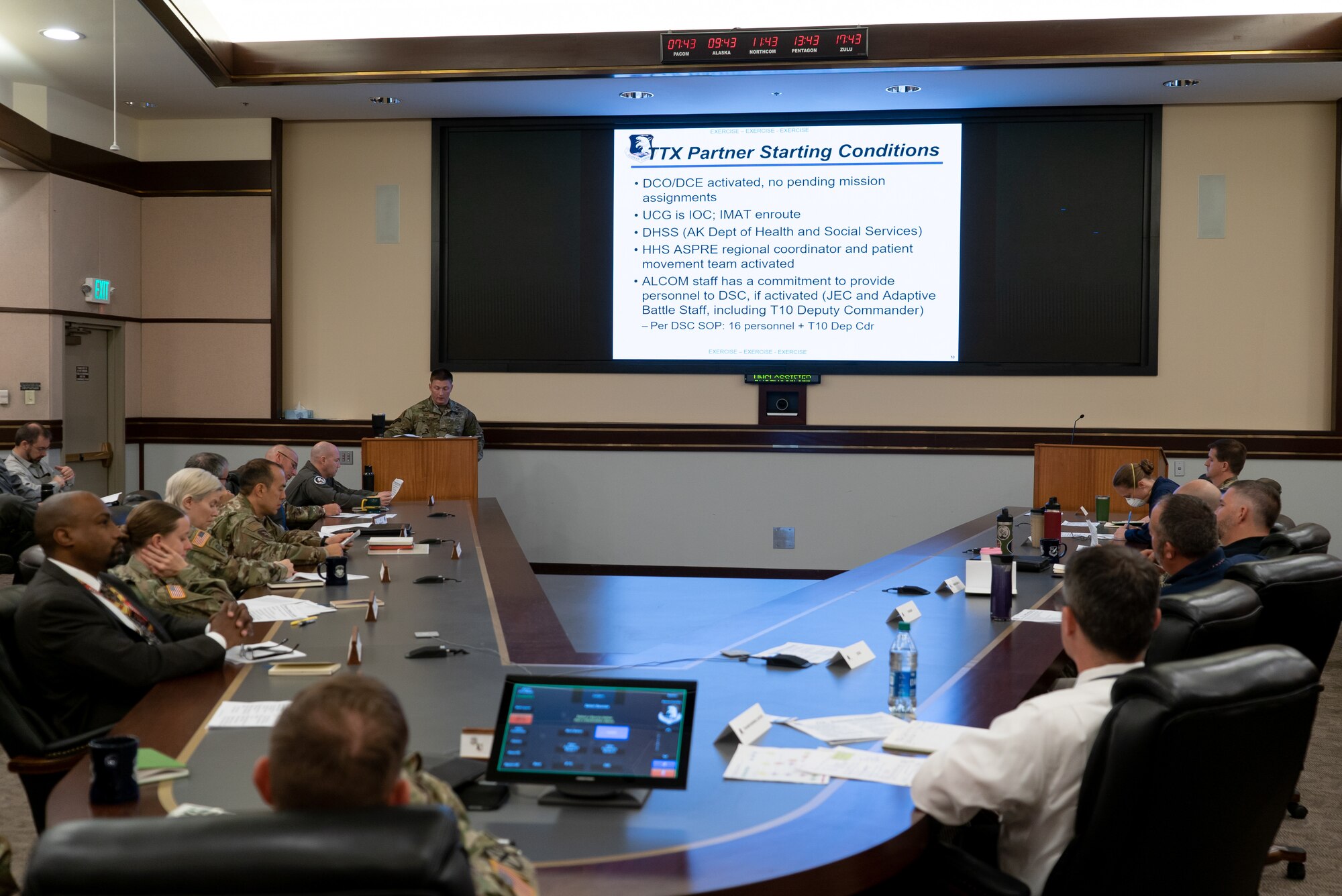A large group of individuals sit at a conference table while a slideshow is projected onto a large screen in front of them