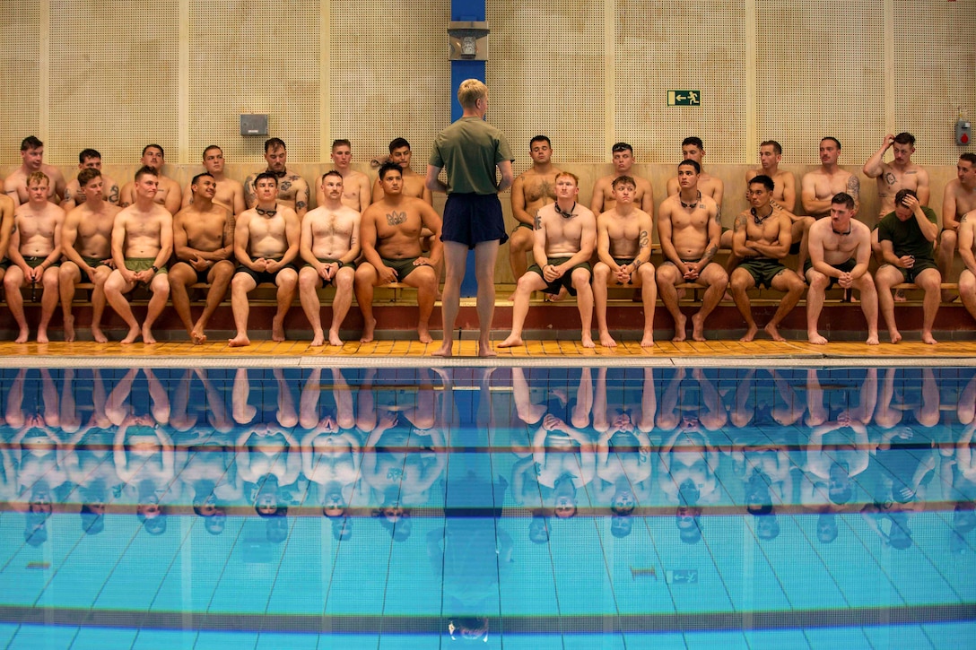 A Marine stands in front of U.S. Marines sitting on benches facing a pool.