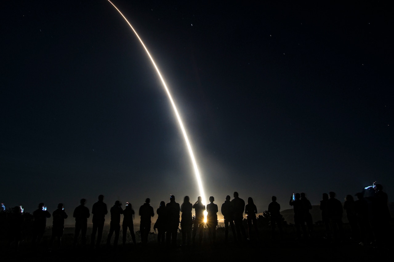 People stand in line holding up their phones as a missile launches in the dark.