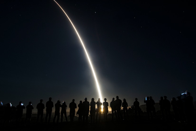 People stand in line holding up their phones as a missile launches in the dark.