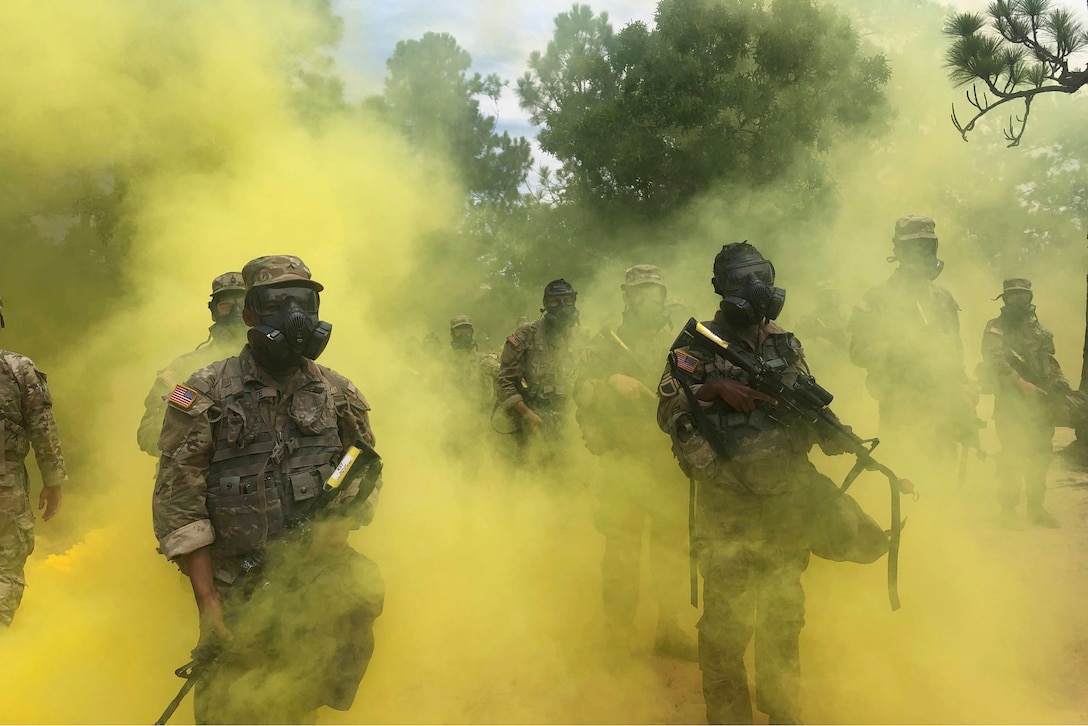 Army basic trainees stand in formation wearing face masks and carrying weapons surrounded by yellow smoke.