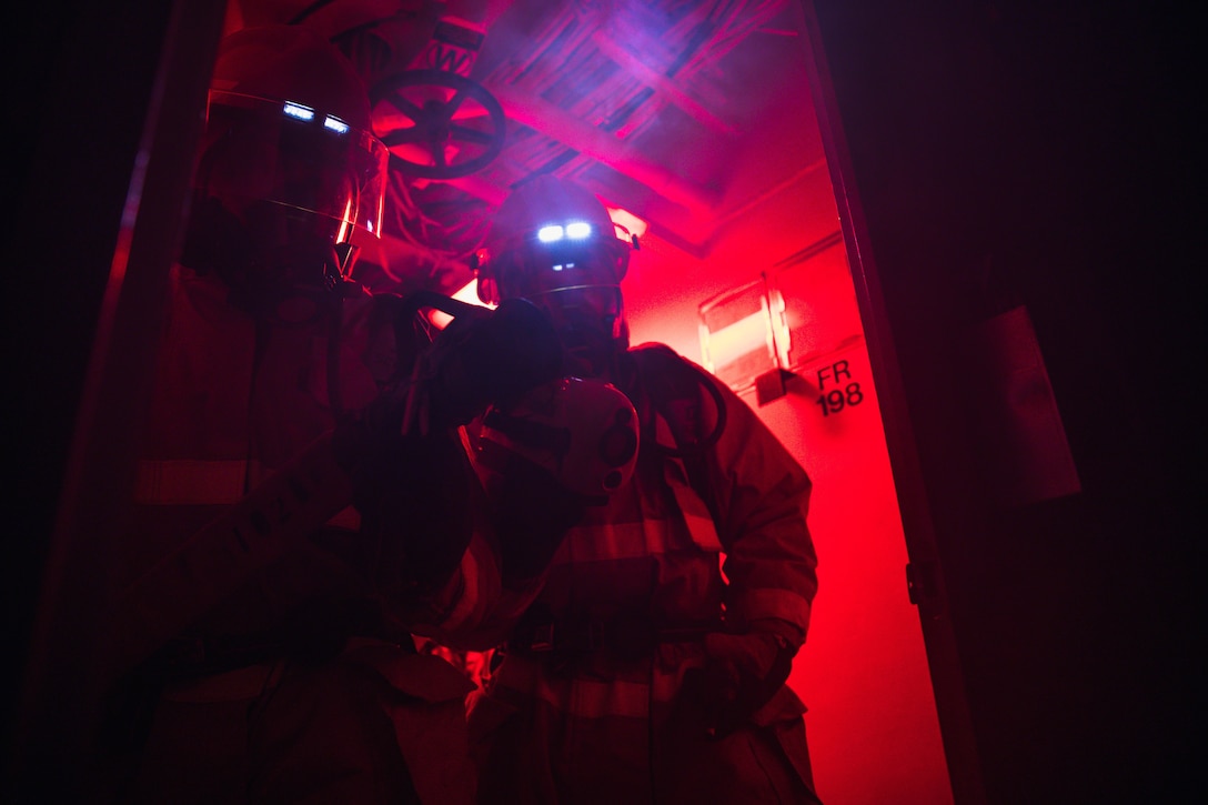 A sailor with lights on his helmet walks through a red lit corridor.