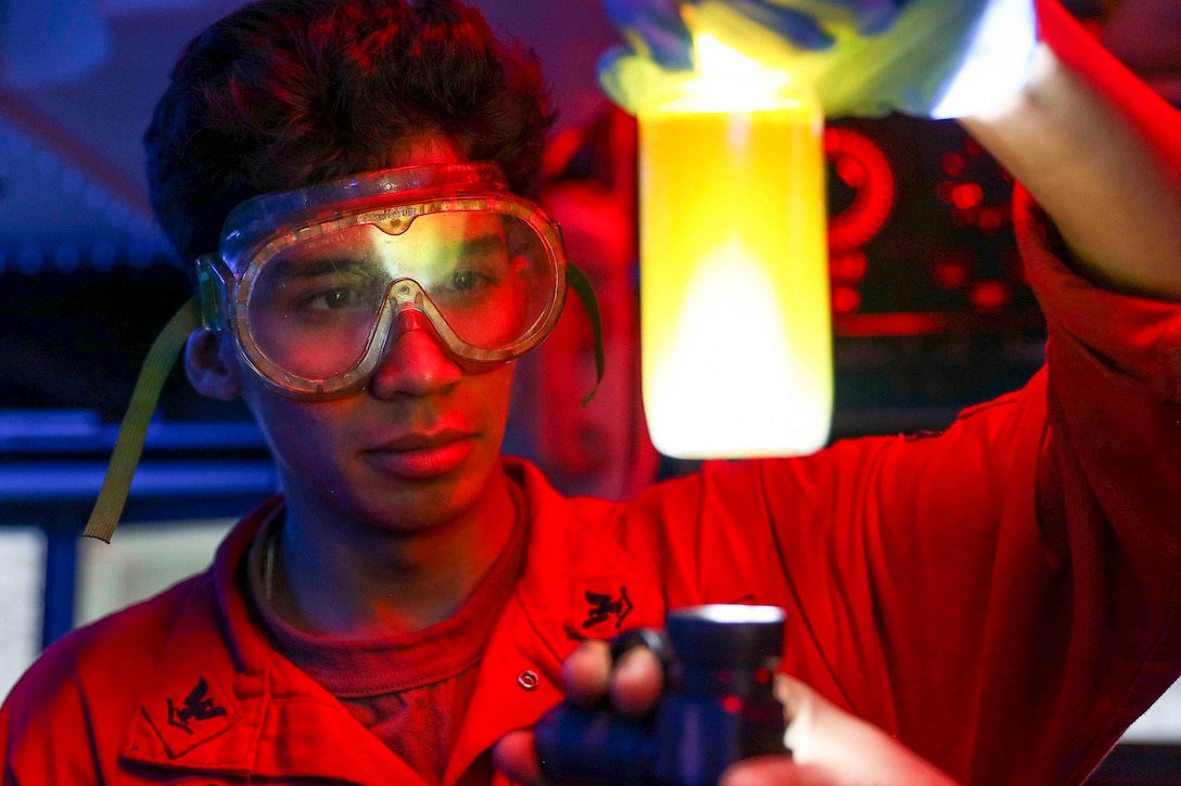 A sailor holds up a bottle of liquid with a light shining on it from below.