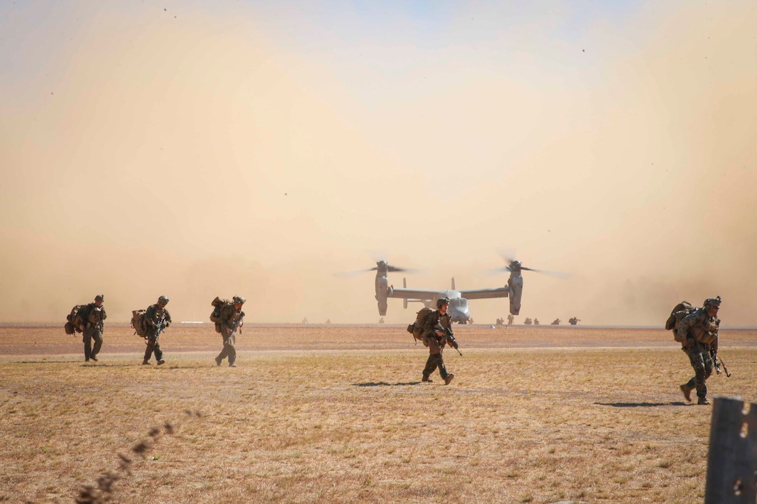 Marines walk across a field as a parked helicopter can be seen in the background.