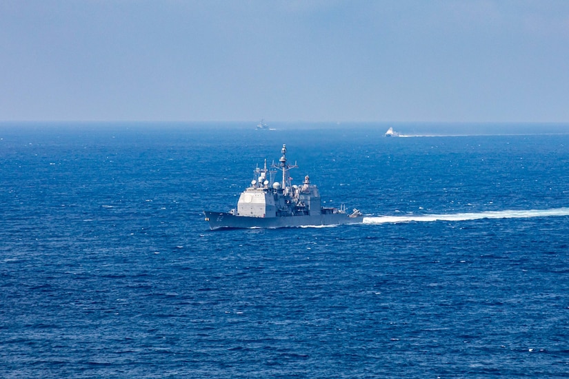 A ship sails through blue water and is followed by two ships in the distance.