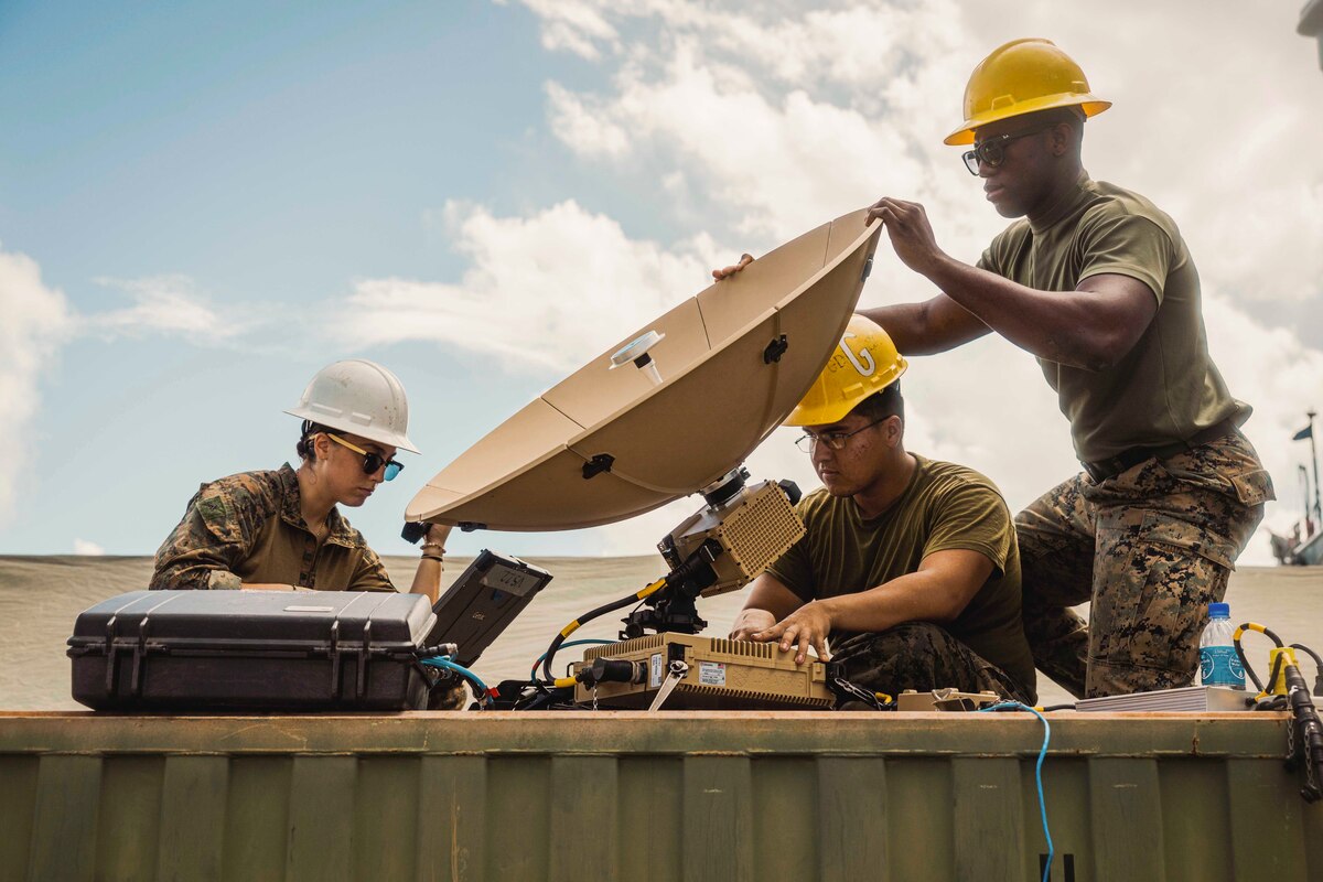 Two service members wearing yellow hard hats position a satellite dish while another service member wearing a white hard hat looks at a laptop.