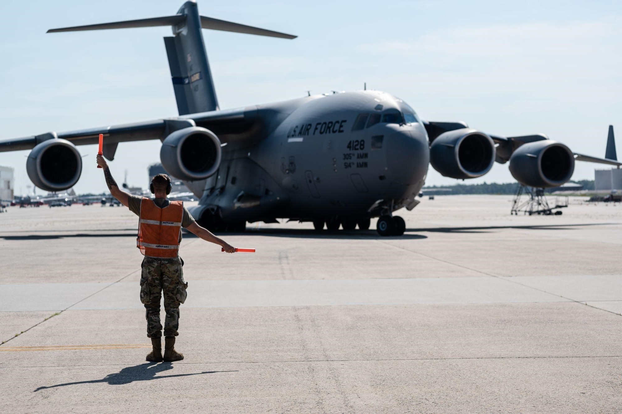 A man wearing a military uniform and a reflective vest directs a plane on a flightline.