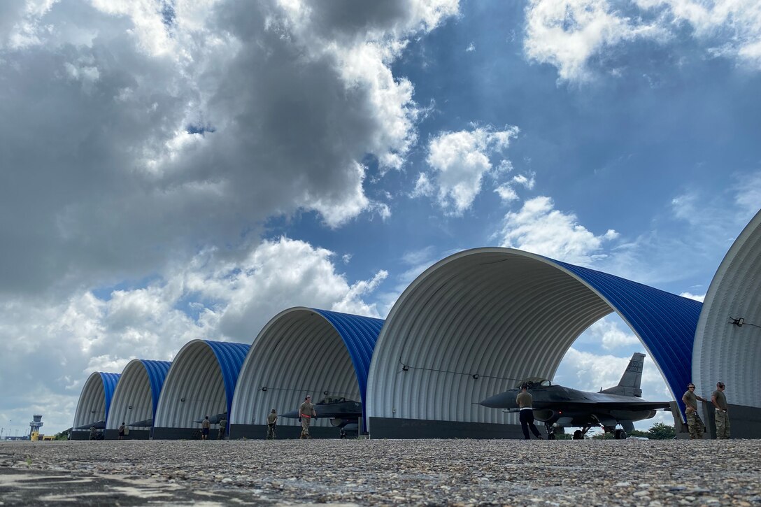 Military jets are parked under oval shelters as airmen walk nearby.