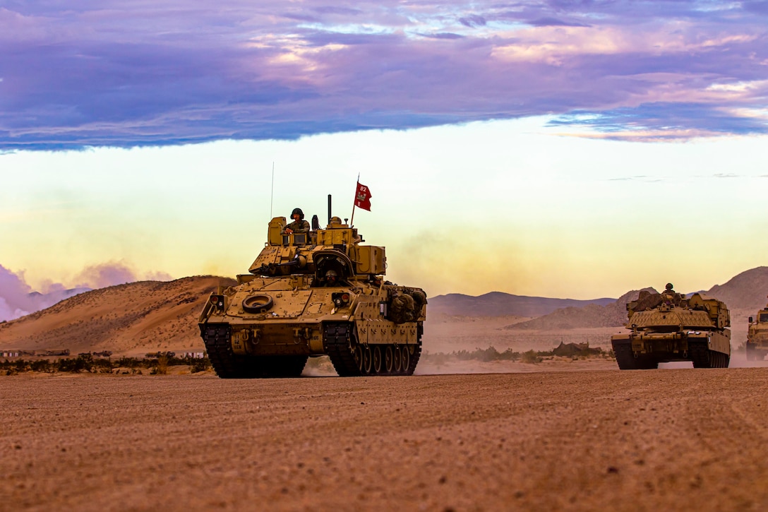 Fighting vehicles drive in formation in a desert.