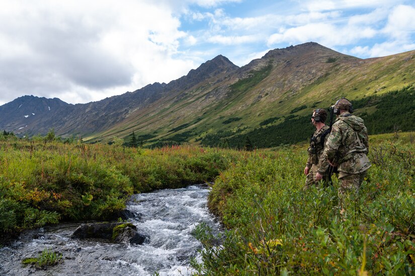Two Airmen stand above a creek observing rugged terrain.