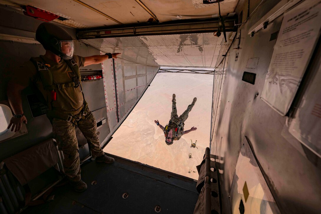 An airman stands at the back of an aircraft as other airmen descend in the sky in a line.