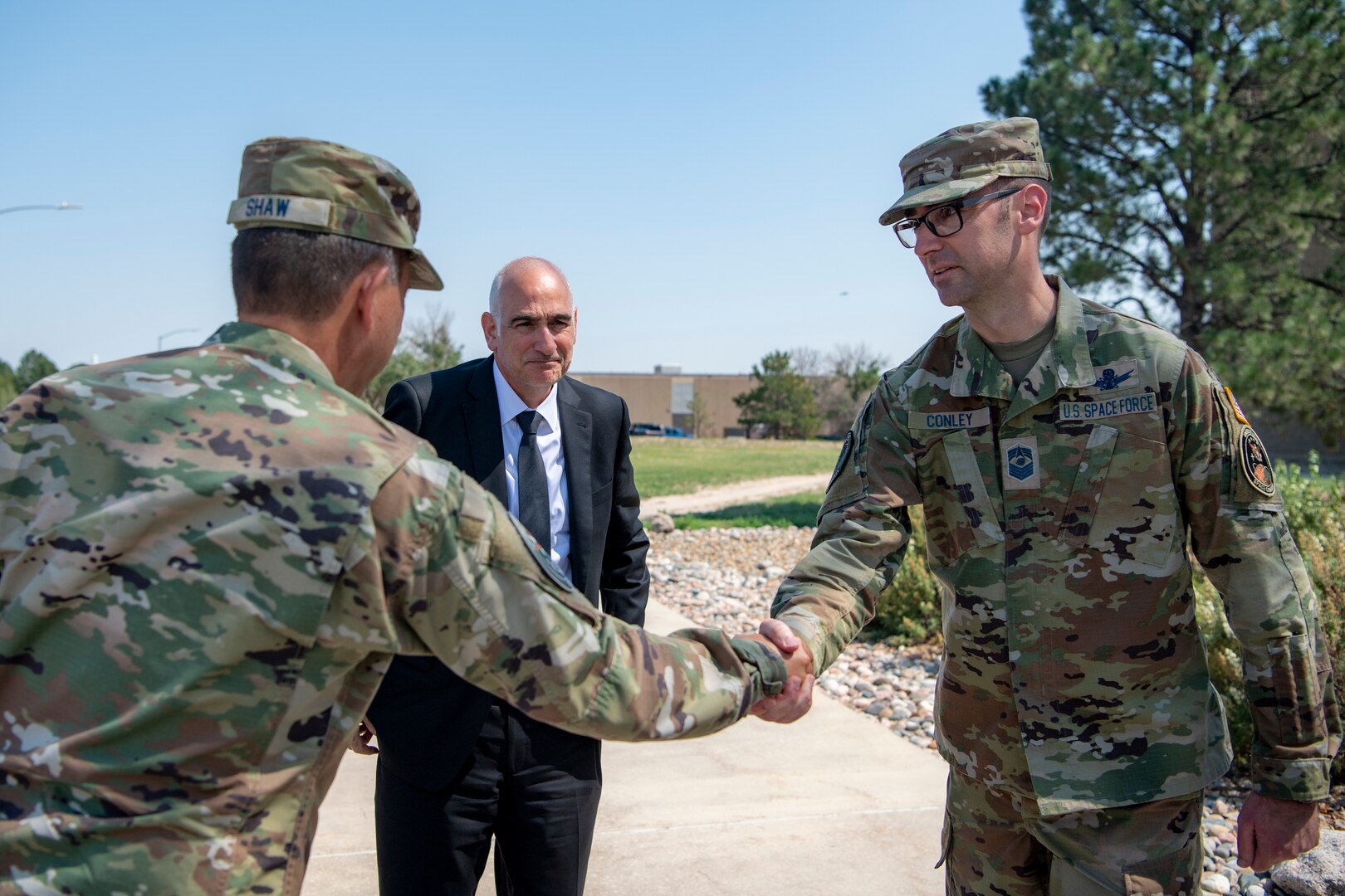 Two military men in uniform shaking hands, man in suit stands behind them