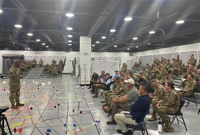 photo of US military sitting in two large groups while one US military member is standing talking to them.