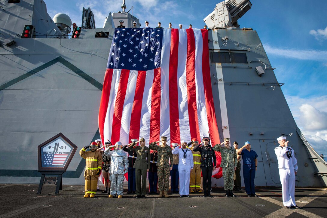 A group of sailors and Marines salute next to a 9/11 memorial on the deck of a ship.