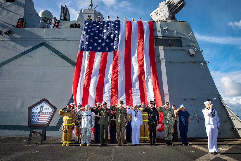 A group of sailors and Marines salute next to a 9/11 memorial on the deck of a ship.