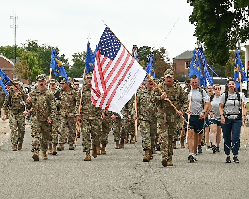 Group of approximately 50 people in military uniform, carrying large American flag and unit pennants, march down street.