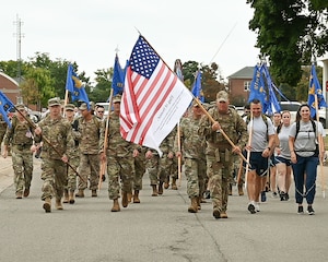 Group of approximately 50 people in military uniform, carrying large American flag and unit pennants, march down street.