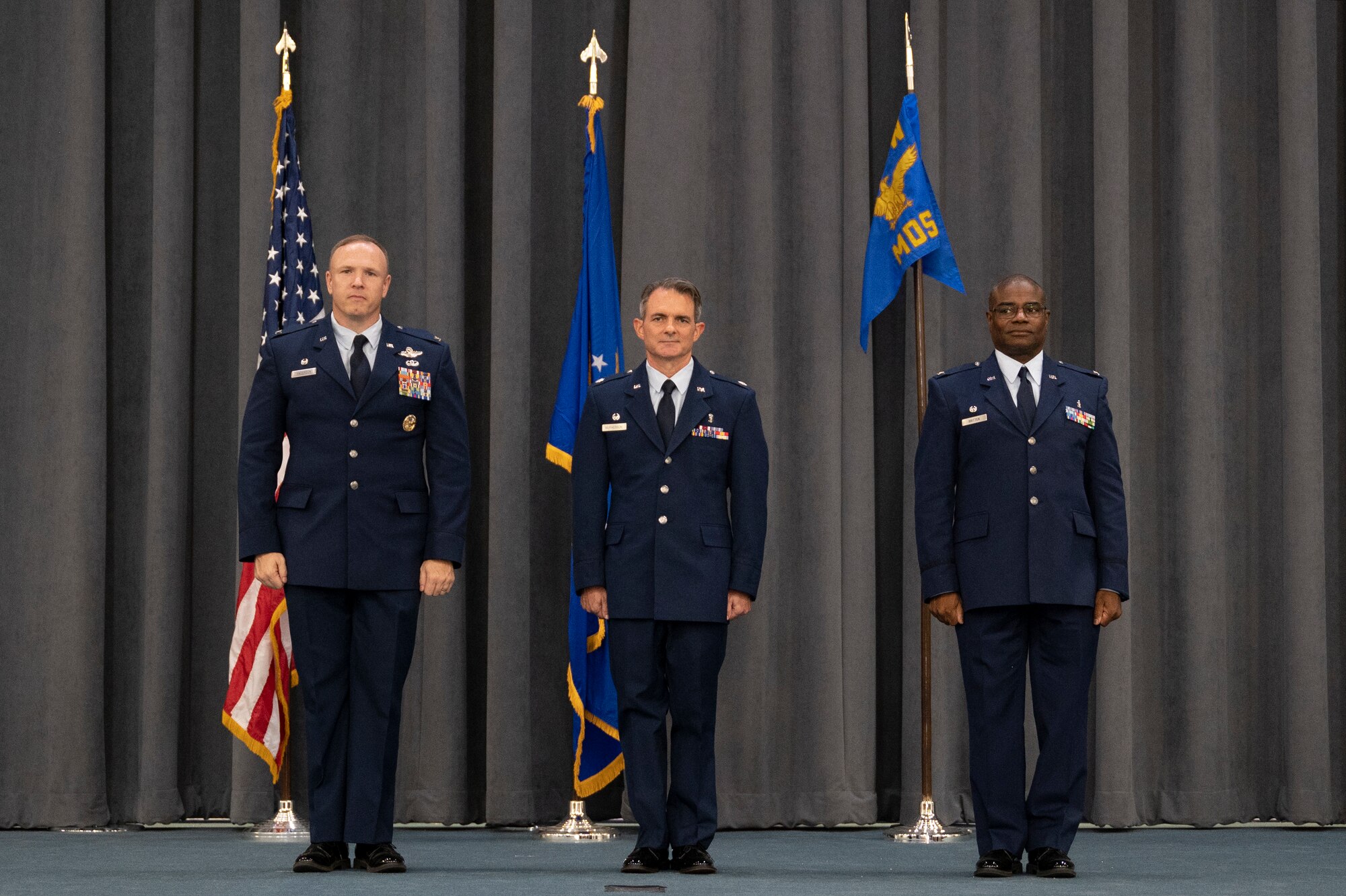 Three men in uniforms standing before flags on a stage.