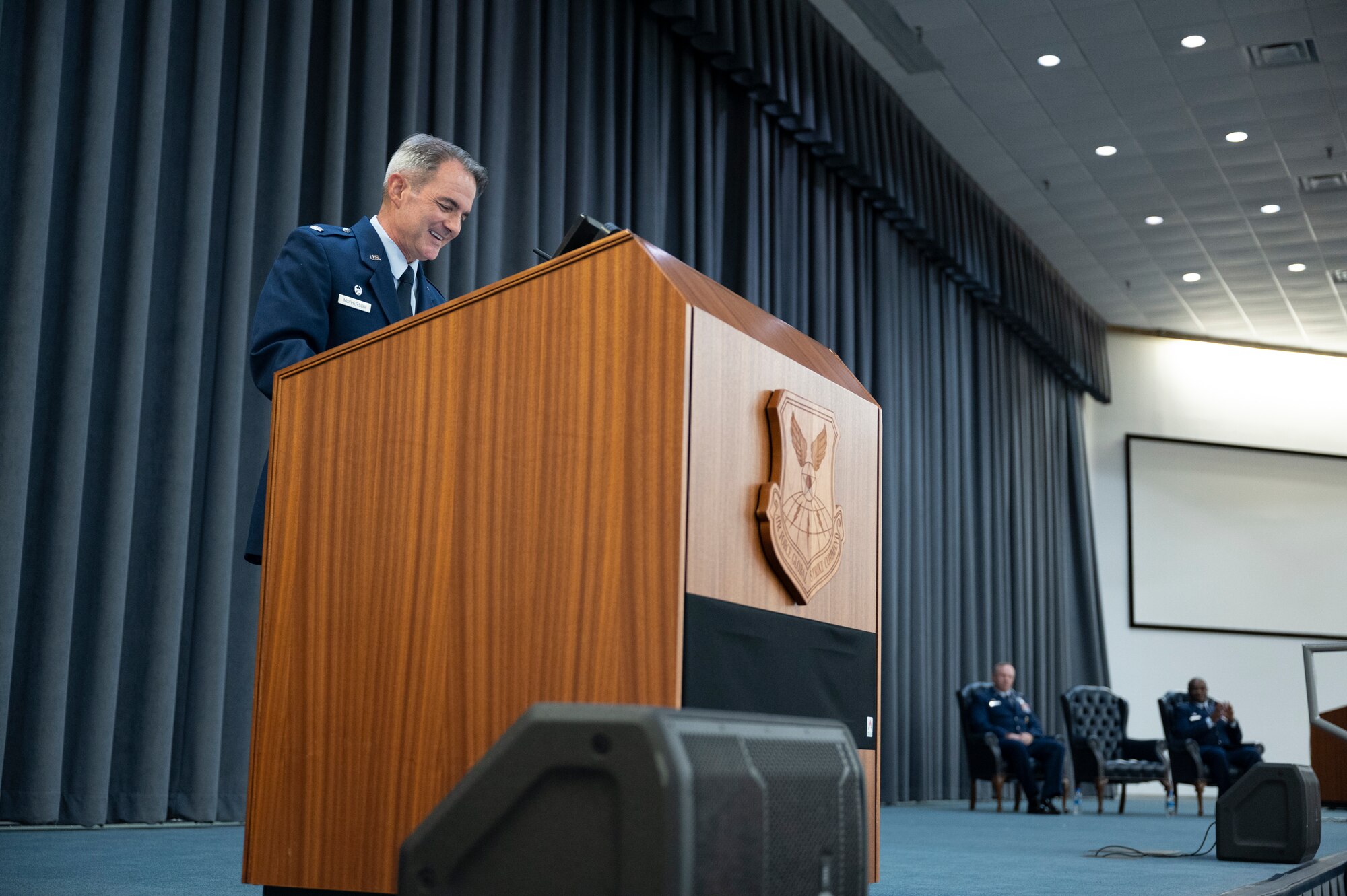 A man in uniform stands at a podium smiling. Two people sit in chairs in the background.