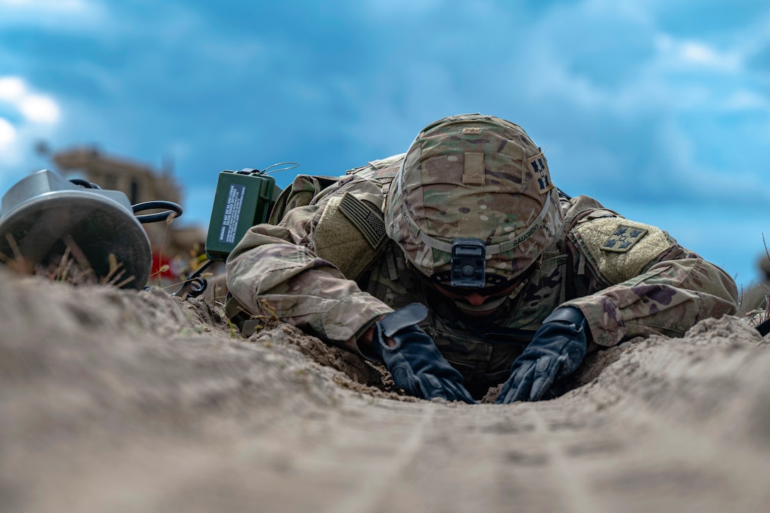 A soldier lies prone and inspects the ground for a possible explosive.