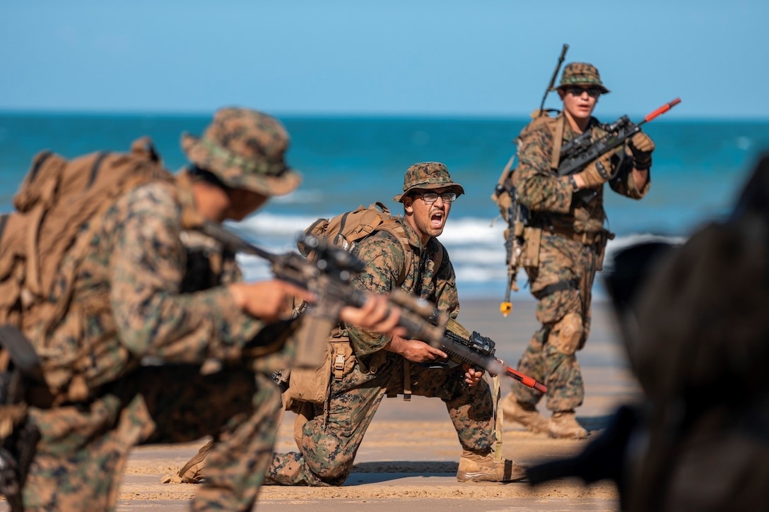 A Marine Corps kneels and yells on a beach as others stand nearby.