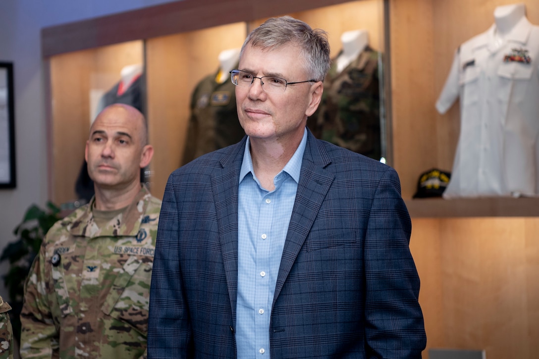 Man in suit wearing glasses standing next to a man in a military uniform, both listening to a briefing