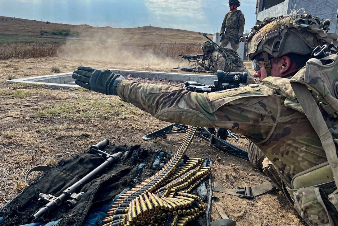 A soldier on the ground looks down the scope of a rifle.